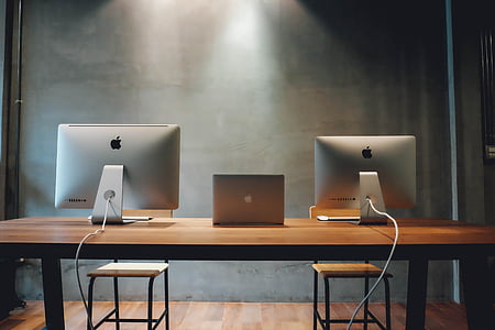 two silver iMac's and silver MacBook in between on brown table thumbnail