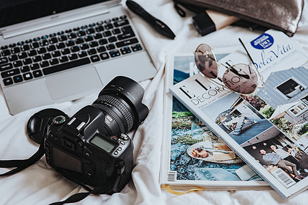 Silver laptop, a camera, magazines and other items on white bed sheets thumbnail