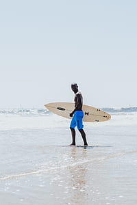 man carrying surfboard while walking on seashore during daytime thumbnail