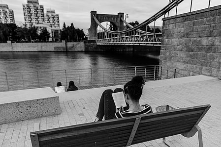 woman sitting on bench while reading book in front of body of water grayscale photo thumbnail