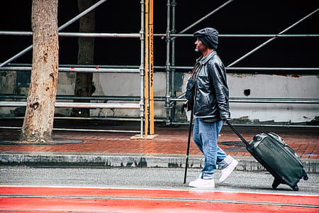 Walking Man in Black Leather Jacket and Blue Denim Pants Holding Luggage Bag thumbnail