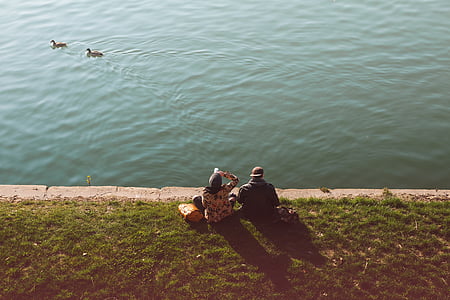 two person sitting near body of water viewing two ducks thumbnail