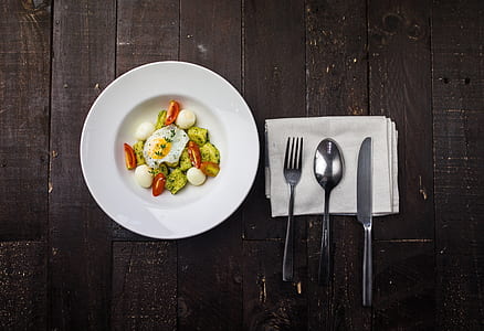 fried egg with greens on round white ceramic plate beside spoon, fork, and knife on top of white linen thumbnail