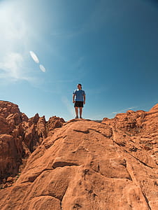 Man Wearing Blue Shirt and Black Shorts Standing on Top of Brown Rock Formations Under Clear Sky thumbnail