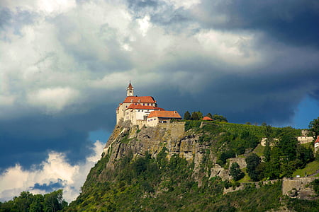 white and orange concrete building on green and grey rocky hill under cloudy sky thumbnail