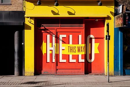 red metal gate hello this way printed on yellow building at daytime thumbnail