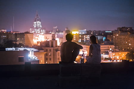 selective focus photography of man and woman sitting on roof top of building looking at cityscape thumbnail