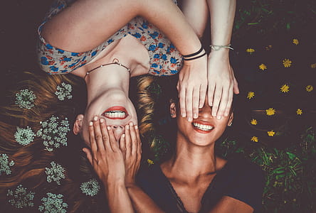 two women leaning on green grass covering both eyes while laughing thumbnail