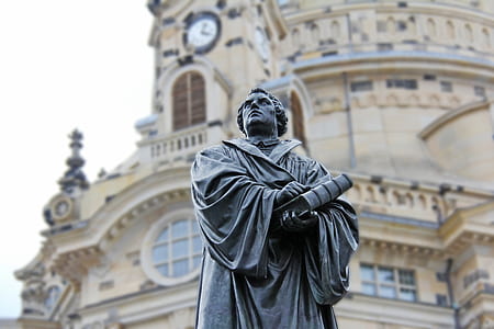 Gray Concrete Statue of Man Holding Book Beside Brown Building thumbnail