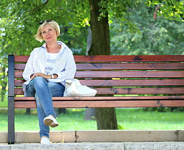 Thinking Woman in White Jacket and White Scoop Neck Shirt Blue Denim Jeans Sitting on Brown Wooden Bench Beside Green Trees during Daytime thumbnail