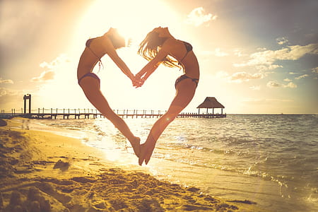 two women jumping while forming heart shape beside seashore during daytime thumbnail