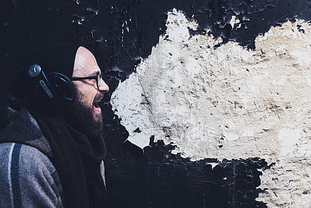 man wearing black headpohones, gray shirt, and black scarf standing beside black and white painted wall thumbnail