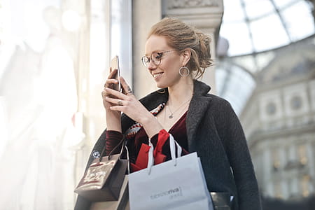A young blond woman wearing a satin dress and an overcoat looks at her mobile phone while holding shopping bags in her hands thumbnail