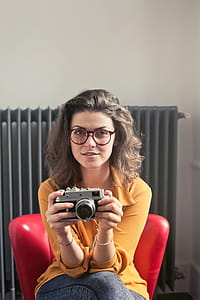 Young Woman Wearing Yellow Collared Shirt And Jeans With Spectacles Holding A Camera While Sitting at Red Chair thumbnail