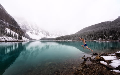 man jumping on calm body of water beside snow capped mountain during daytime thumbnail