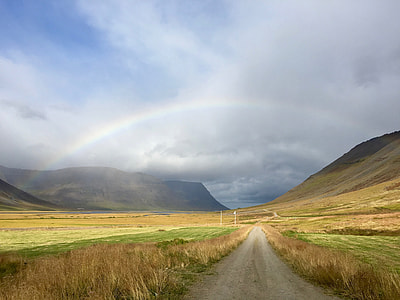 gray road between green grass near mountain under gray clouds with rainbow at daytime thumbnail