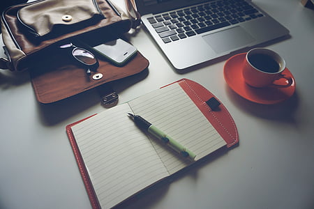 white pen on white notebook beside the orange ceramic coffee cup with saucer located on top of white table thumbnail