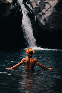 man in body of water wearing brown cowboy hat facing waterfalls at daytime thumbnail