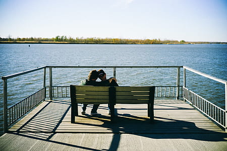 man and woman sitting down on bench in front of body of water during daytime thumbnail
