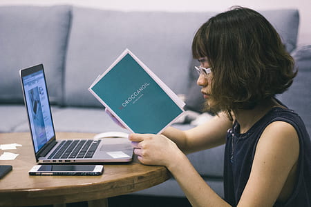 woman in black tank top beside coffee table with laptop computer reads book thumbnail