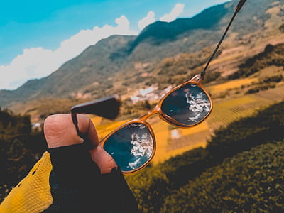 person holding black lens sunglasses with gold frames with reflection on white clouds in sky thumbnail