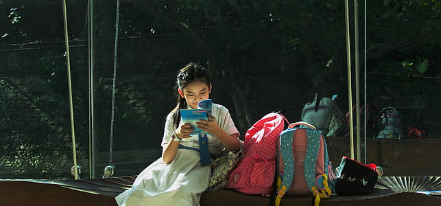 girl wearing white dress reading book beside two backpacks during daytime thumbnail