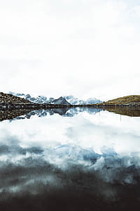 calm lake towards snow covered mountains under white sky thumbnail