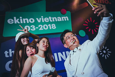 Man in White Suit Jacket Holding Phone in Front of Three Women thumbnail