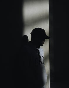 silhouette photo of man wearing gray GAP hoodie and black cap leaning on wall thumbnail