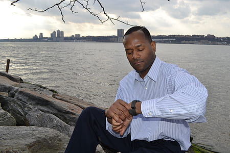 man looking at his watch while sitting on rock near body of water thumbnail