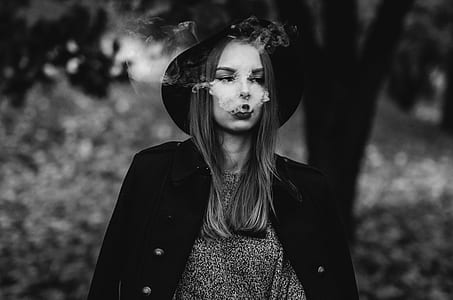 greyscale photo of woman wearing hat and jacket blowing smoke thumbnail