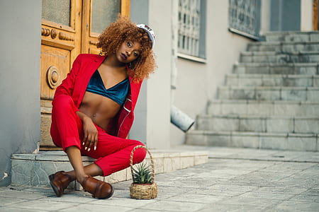 Woman Wearing Red Coat and Pants Sitting Outside Gray Building thumbnail
