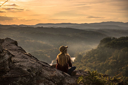 A woman wearing a hat sits on area with mountains thumbnail