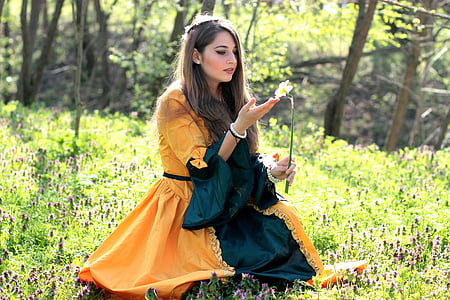 woman wears orange and blue dress sits on green grass field holds petaled flower at daytime thumbnail