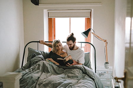man sitting on bed beside woman reading a book inside white painted room thumbnail