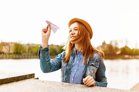 Woman in Blue Denim Button-up Jacket and Brown Hat About to Fly a Paper Plane Beside a Body of Water thumbnail