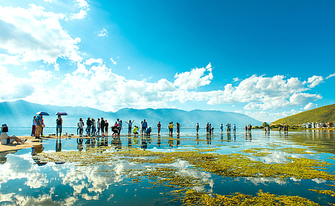 group of people standing on shallow body of water near mountains under cloudy blue sky thumbnail