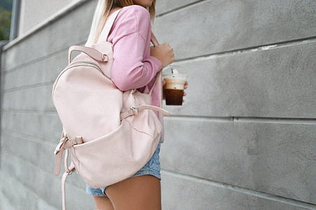 woman with beige leather backpack standing beside gray concrete wall holding plastic disposable cup on focus photo thumbnail