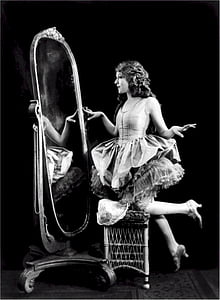 grayscale photography of woman in white sleeveless dress staring her self in vanity mirror thumbnail