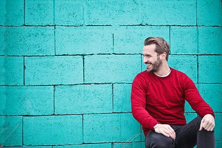 Young Adult Man wearing a red sweater leaning on a turquoise brick wall thumbnail