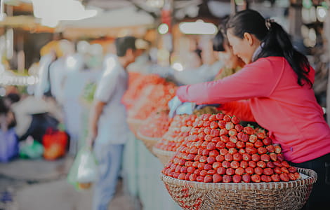 Selective Focus Photography Of Woman Wearing Red Hoodie Standing Behind Of Basket Full Of Fruits thumbnail