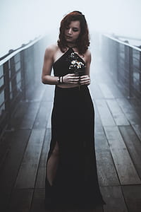 woman wearing black dress holding white petaled flower standing on wooden dock thumbnail