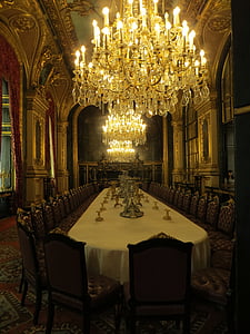 three lighted crystal chandeliers over long dining table with empty chairs inside dining hall thumbnail
