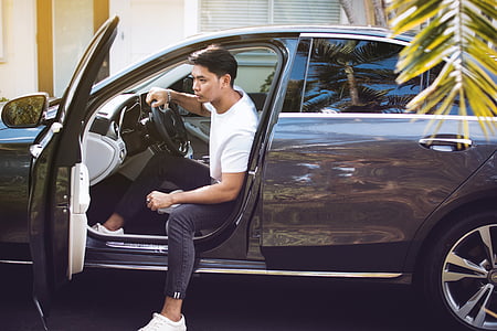 man sitting on drivers seat of car near white building thumbnail
