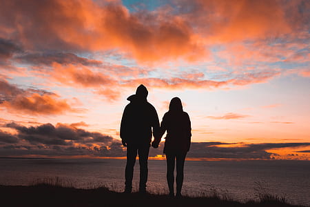 silhouette of man and woman holding hands facing sea at sunset thumbnail