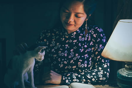 Woman Caressing a Cat While Reading Book Lighted by a Table Lamp in Dark thumbnail