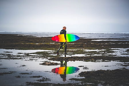 man carrying multicolored surfboard walking on beach thumbnail