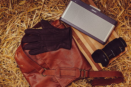 brown leather glove, black zoom lens, brown leather satchel bag, and gray Bose speaker on brown dried leaves thumbnail