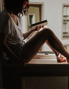 woman seating on window while holding book thumbnail