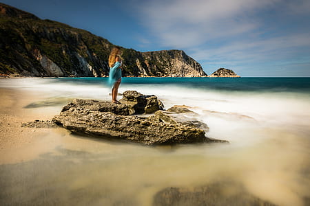 woman in blue shirt standing on top of gray stone beside body ofwater thumbnail